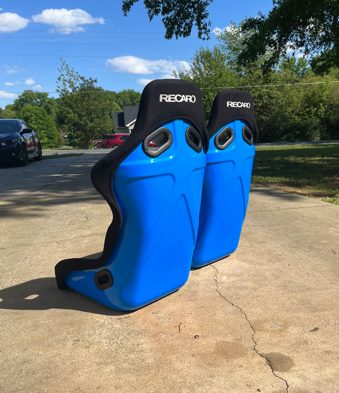 Two blue Recaro racing seats outdoors on a driveway, with trees and parked cars in the background