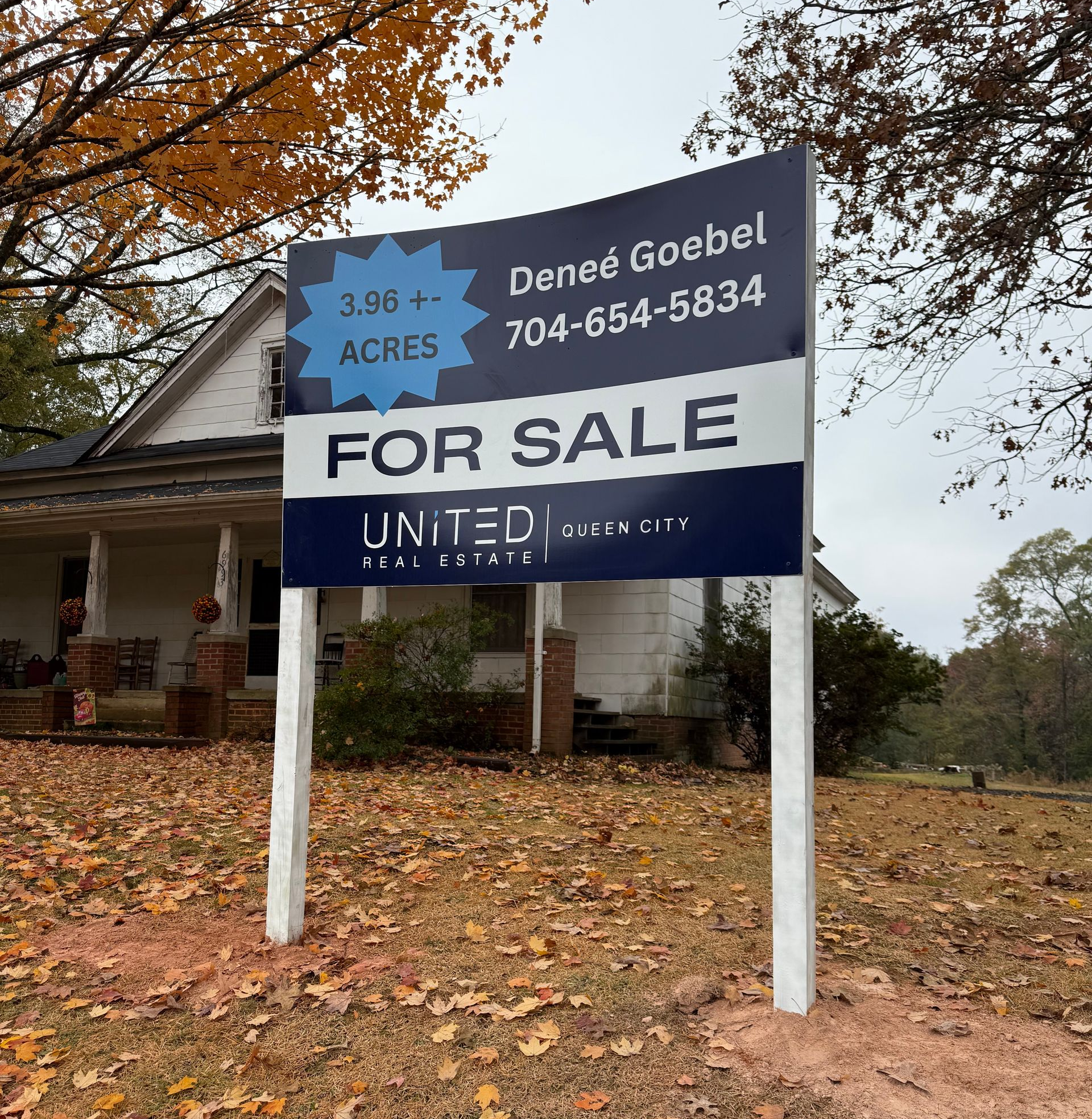 For sale sign in front of a house, with blue and white panels and a phone number, under autumn trees