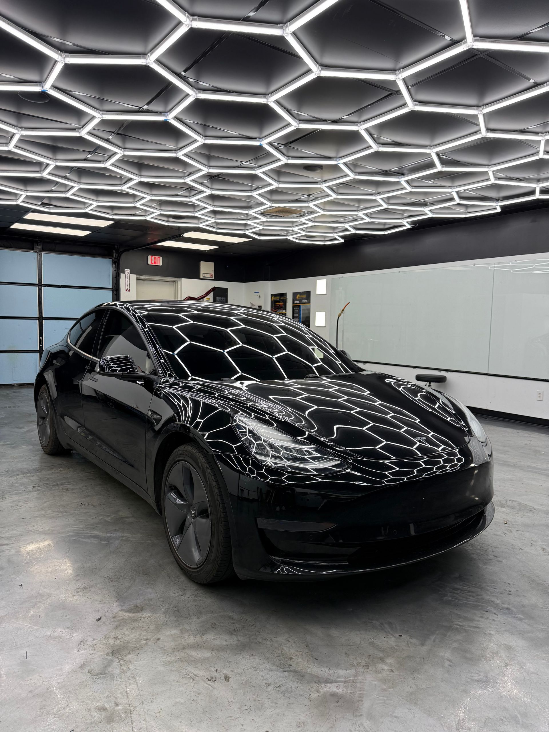 Black Tesla sedan parked in a modern garage with hexagonal ceiling lights reflecting on its glossy surface