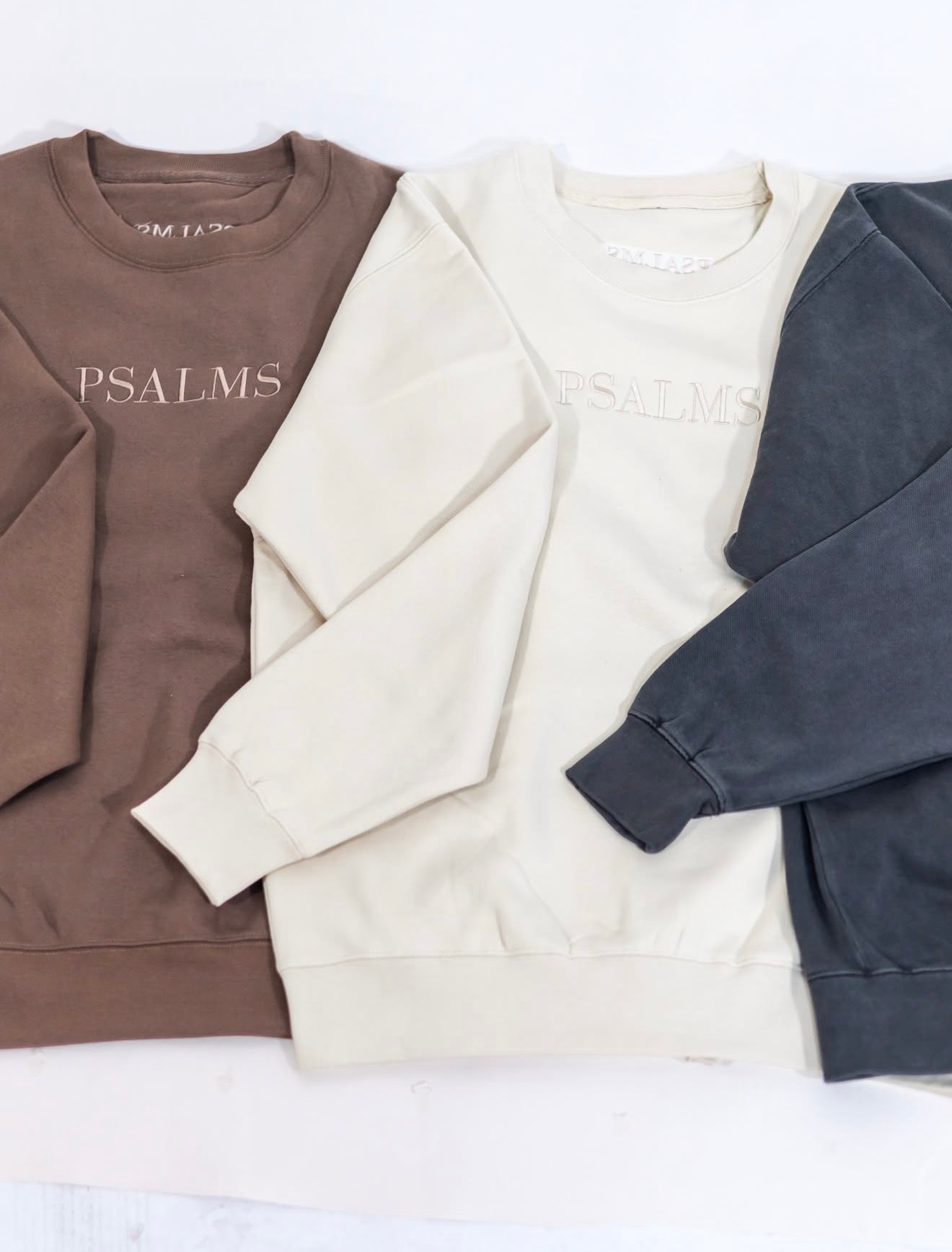 Three crewneck sweatshirts in brown, cream, and slate blue laid side by side