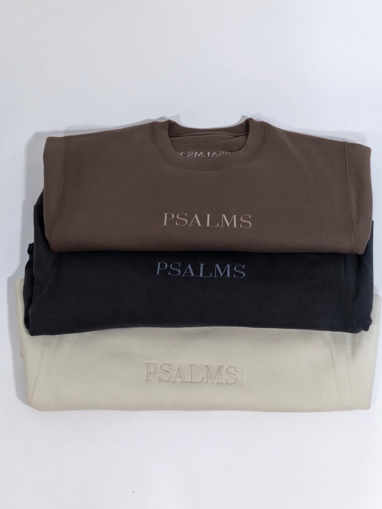 Stacked folded sweatshirts in brown, black, and cream with “PSALMS” printed on each.