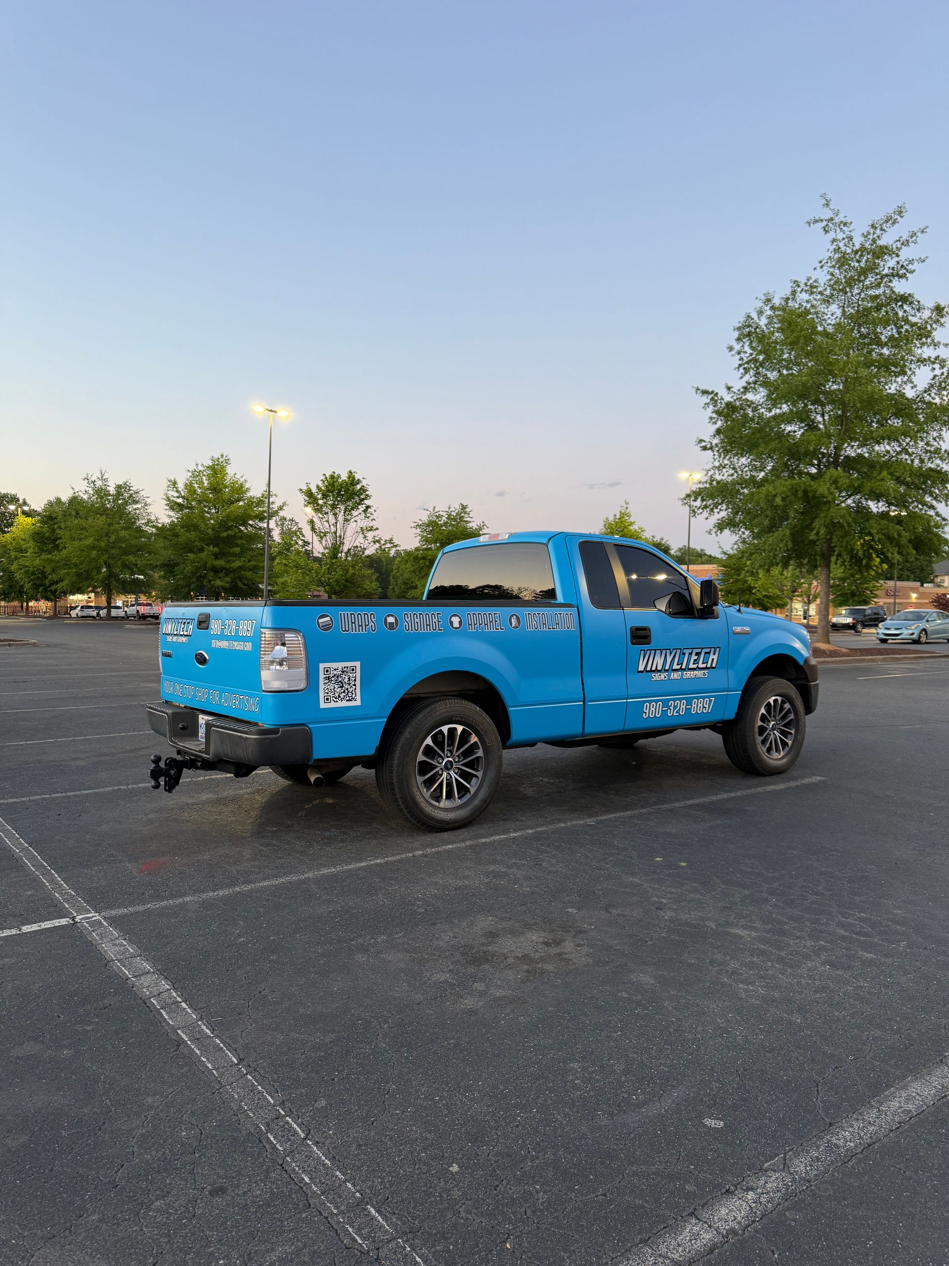 Blue pickup truck parked in an empty lot at dusk, with trees and lights in the background.