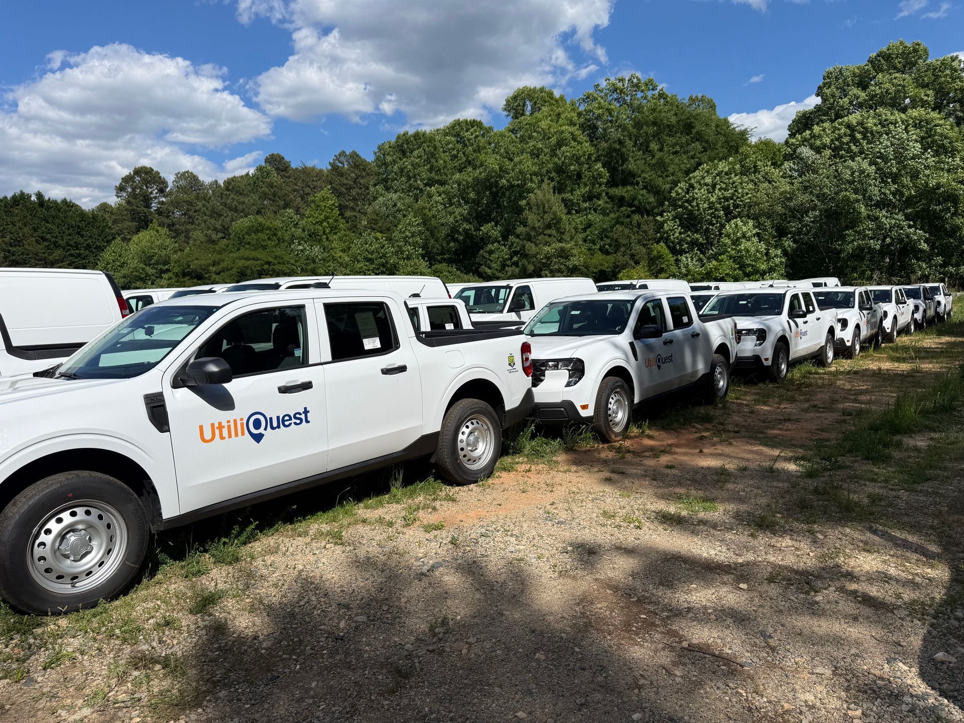Row of white utility trucks parked on grass beside a wooded area under a blue sky
