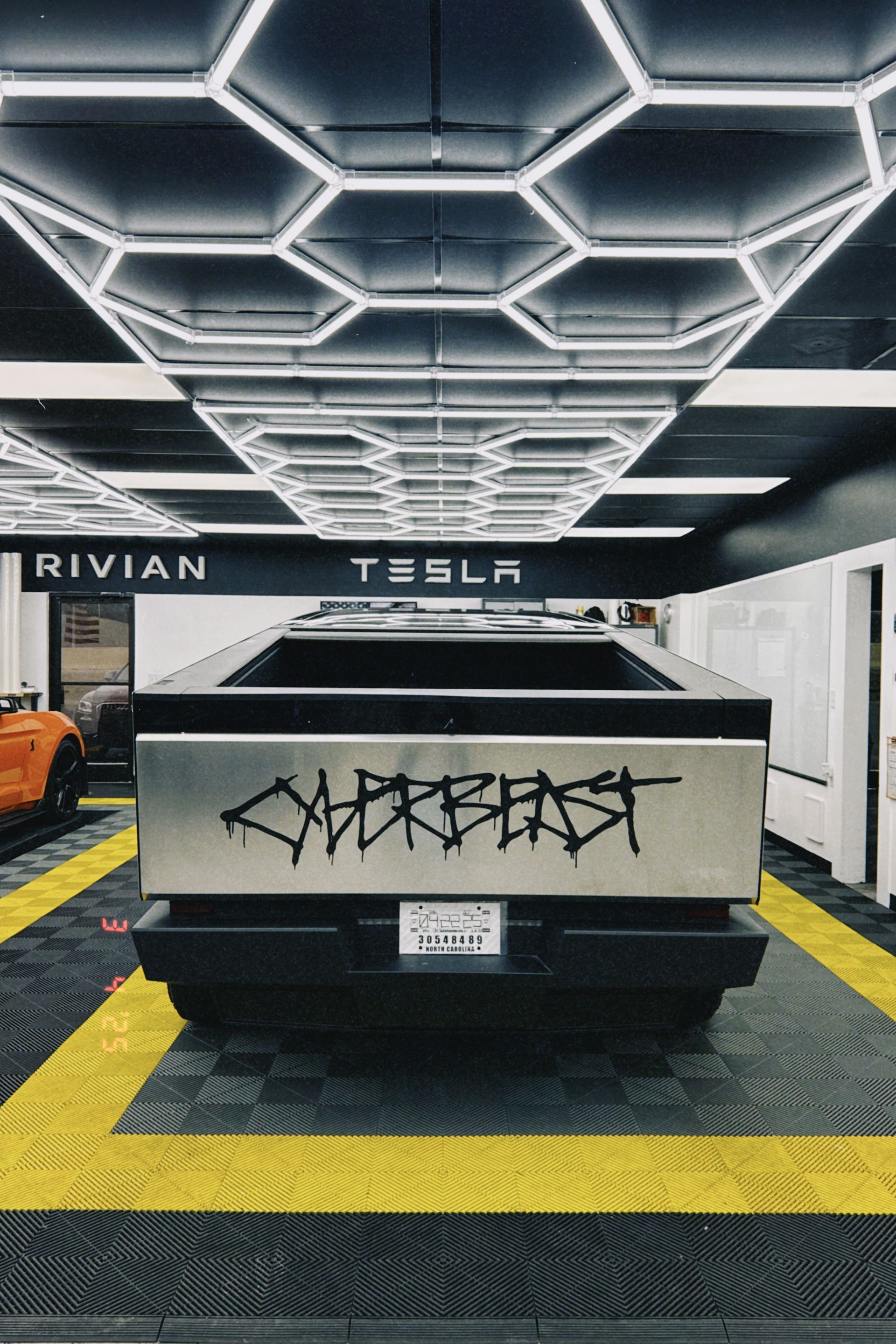 Tesla Cybertruck displayed indoors under hexagon ceiling lights, with Rivian and Tesla signs in background