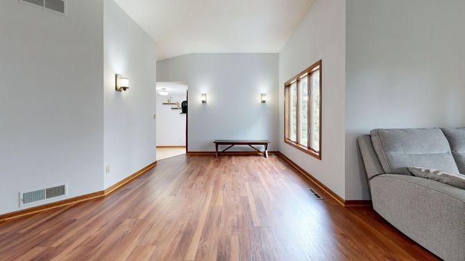 Empty living room with hardwood floors, gray sofa, wall sconces, and a windowed side wall.