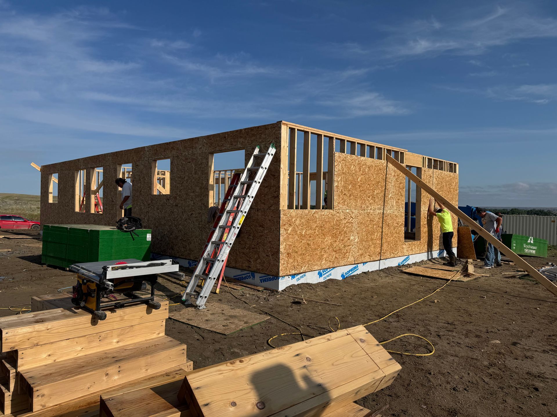 Construction of a wood-framed house on a sunny day; workers, a ladder, and building materials visible.