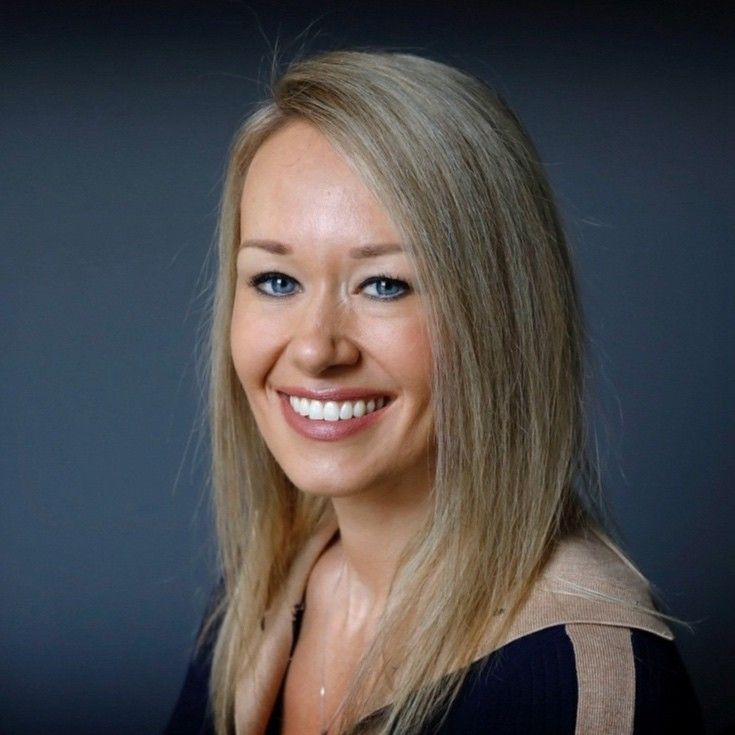 Woman with blonde hair smiles, looking towards the camera; she has blue eyes and is wearing a dark blue and tan top.