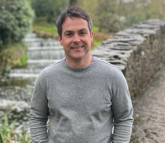 Man in gray sweater smiles outdoors near stone wall and small waterfall.