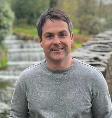Man smiling in a grey sweater; a stone structure and a waterfall are in the background.