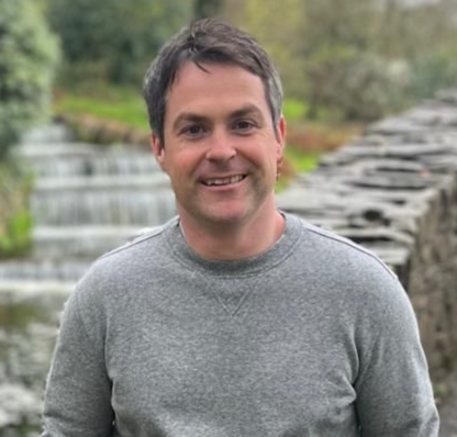 Man smiling outdoors, wearing a gray sweatshirt, with a waterfall in the background.