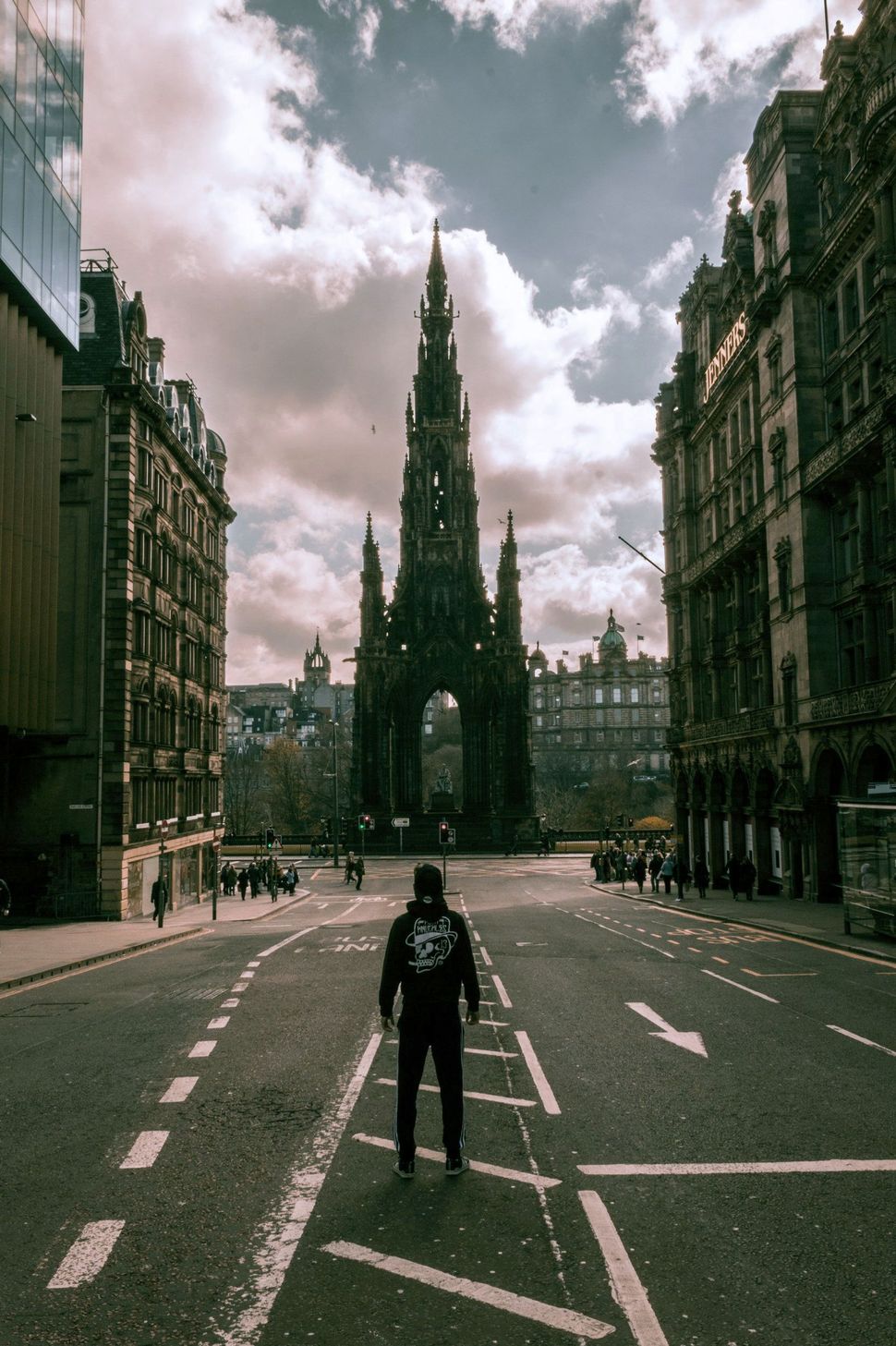 Man standing on street, facing Scott Monument in Edinburgh. Cloudy sky, buildings on either side.