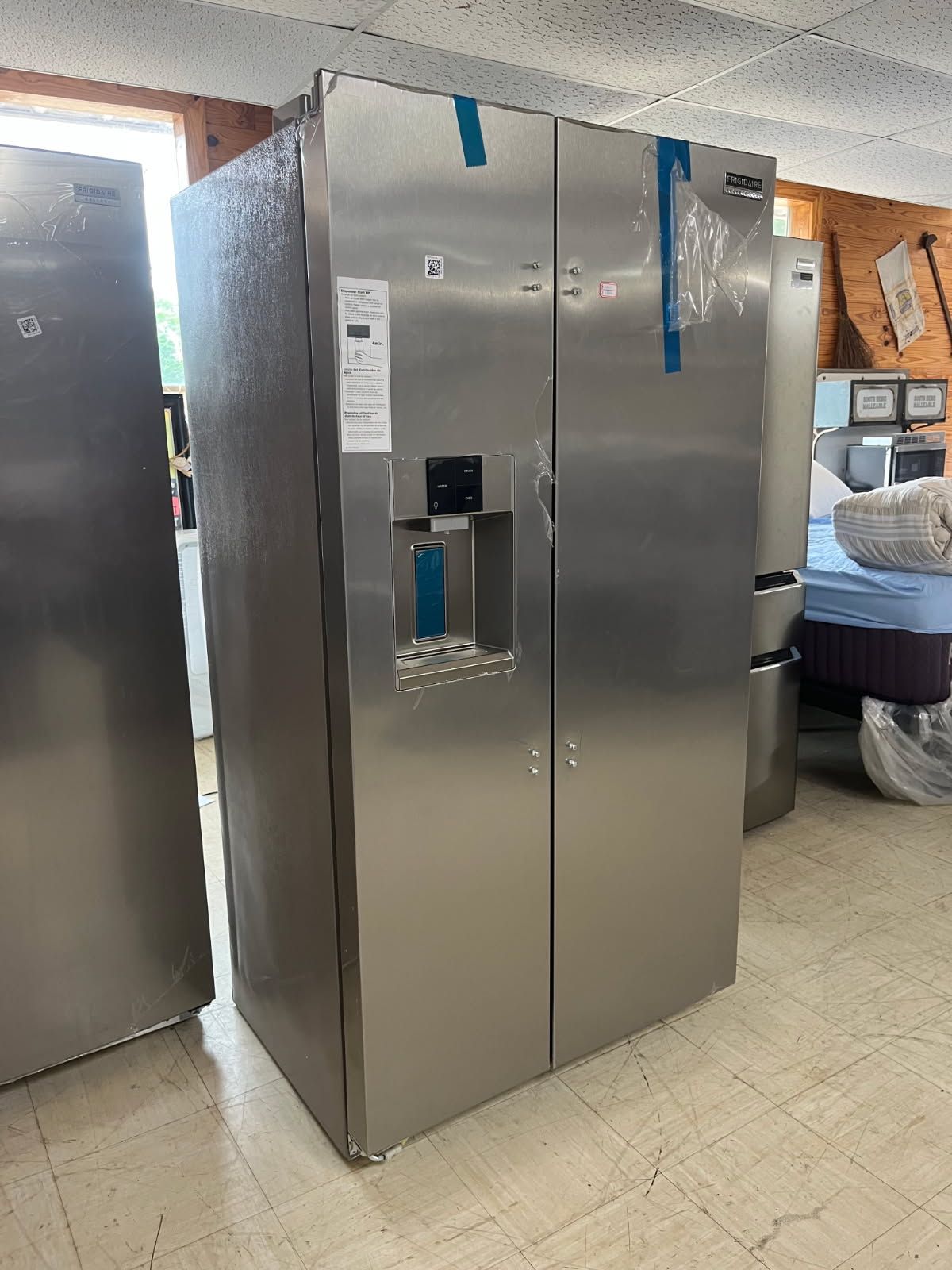 Stainless steel refrigerator in a store aisle, with blue tape on the doors and a water dispenser.