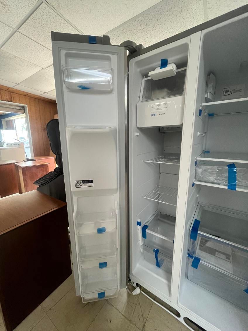 Open refrigerator with shelves, bins, and blue tape in a room; a person is partly visible behind it