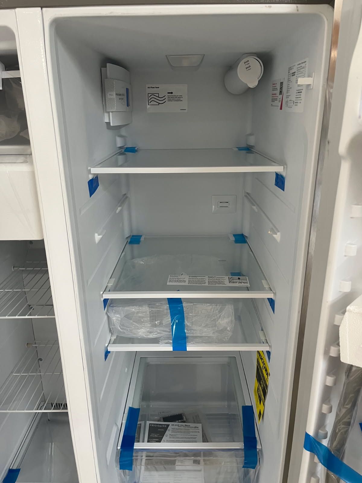 White refrigerator interior with empty wire shelves and blue tape markers, viewed with the door open