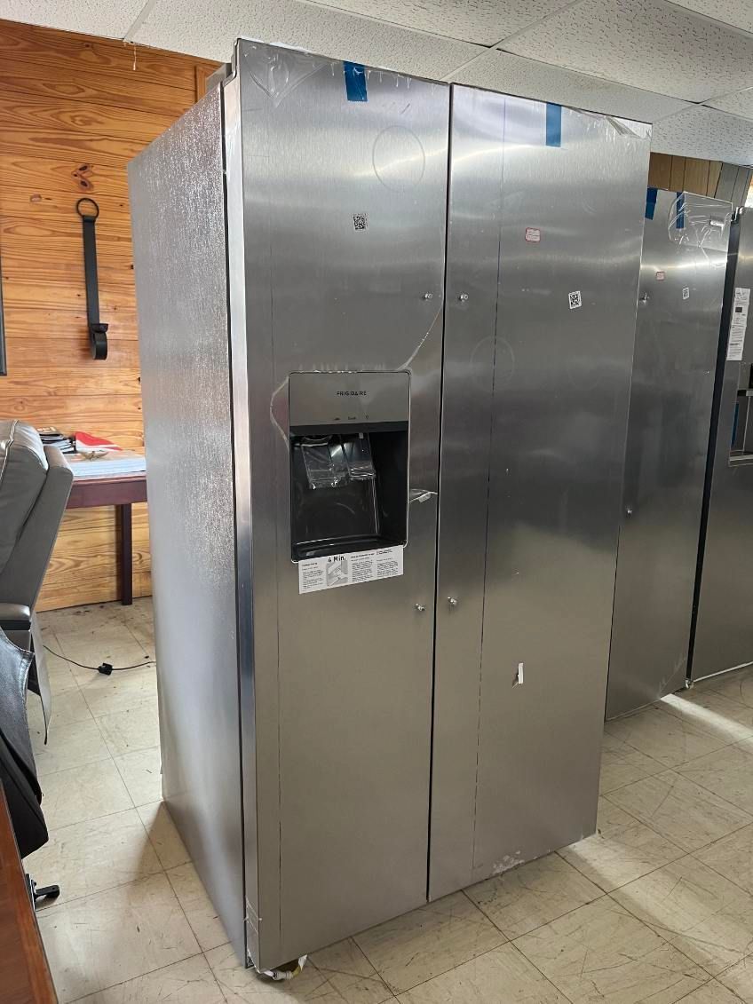 Large stainless steel refrigerator in a room with tiled floor and brick wall, with one door open exposing shelves