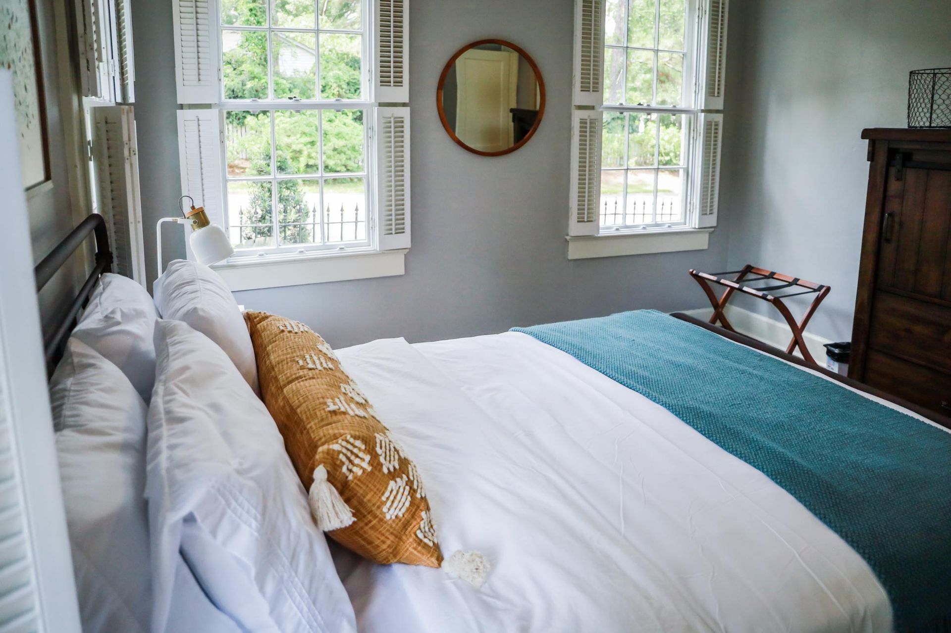 Bright bedroom with a white bed, teal blanket, mustard pillow, and large windows overlooking greenery