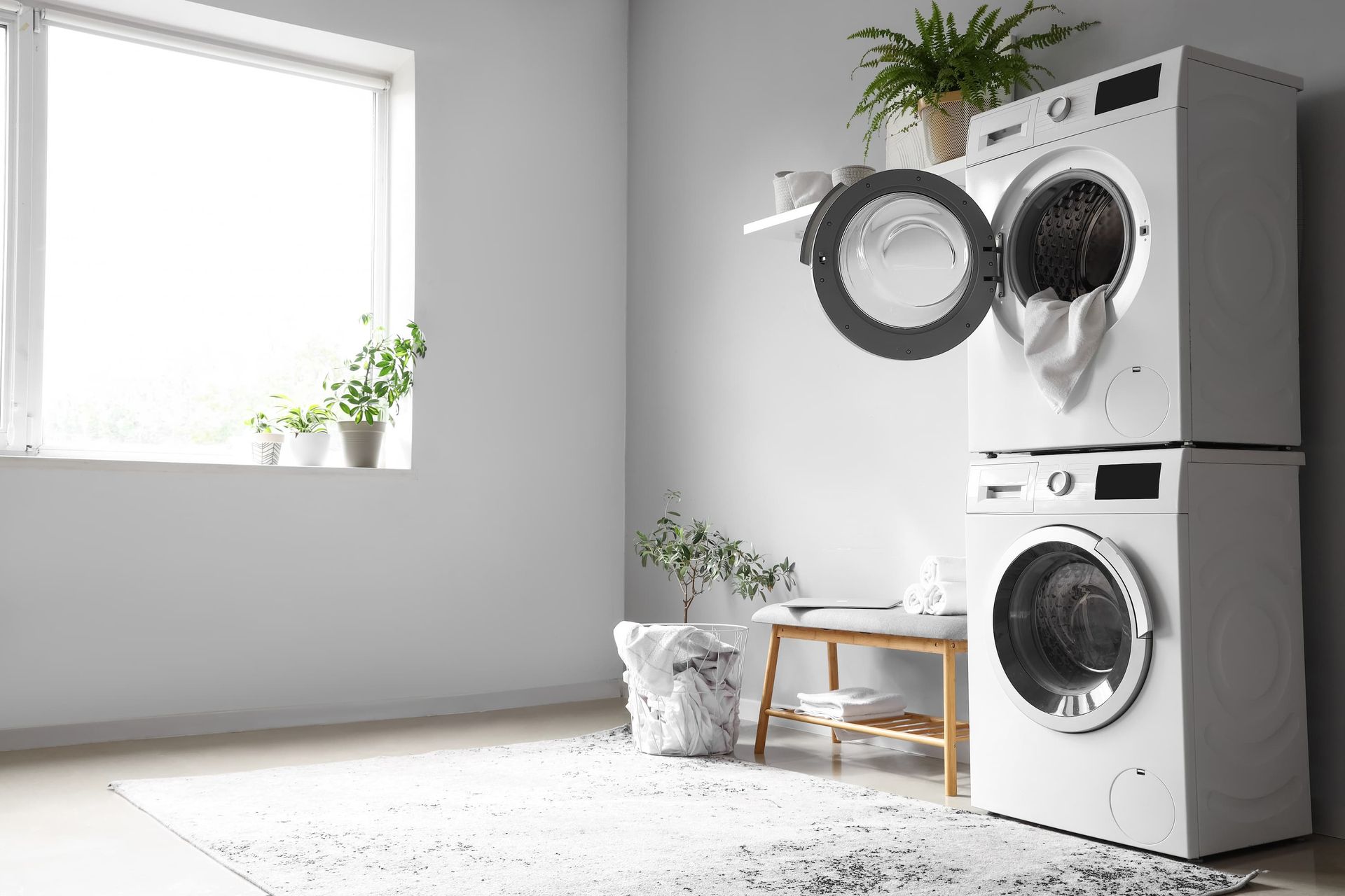 Stacked washer and dryer in a bright laundry room with a window, plants, and a small bench