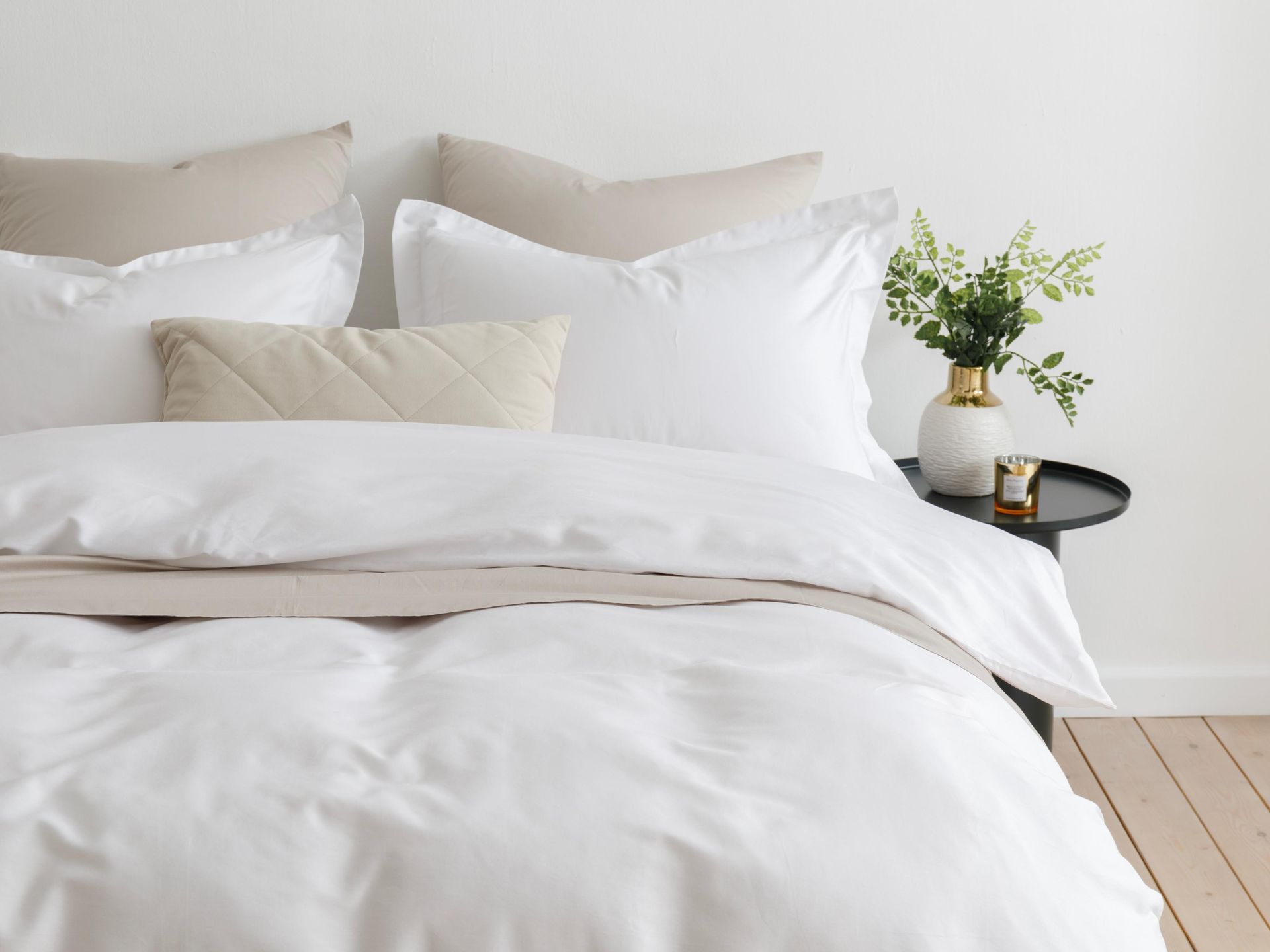 White bed with pillows and rumpled duvet beside a small table with a plant in a bright bedroom