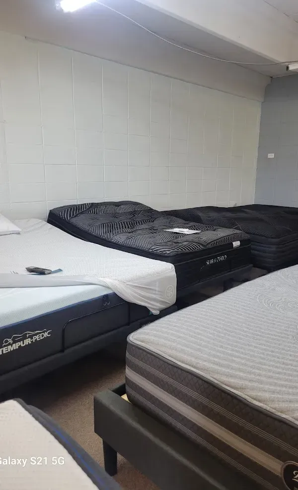 Small room with several mattresses and beds lined against a white wall, one with a dark blanket.