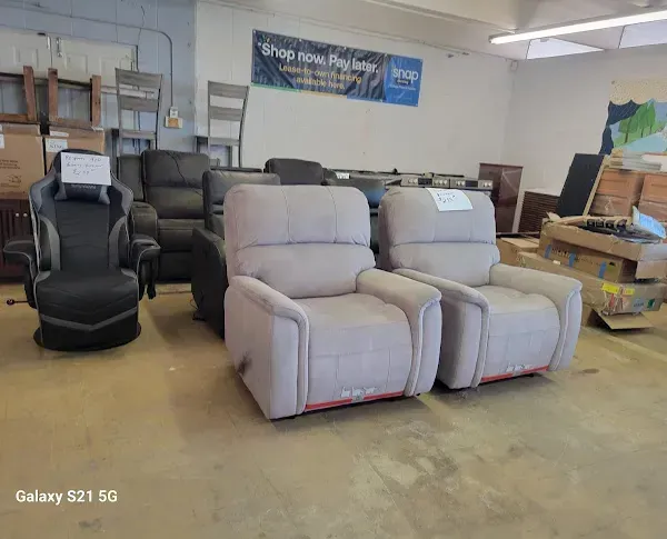 Furniture showroom with two light gray recliners and several darker recliners in a warehouse-like space