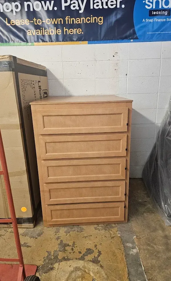 Wooden five-drawer dresser in a warehouse aisle beside a white cinderblock wall.