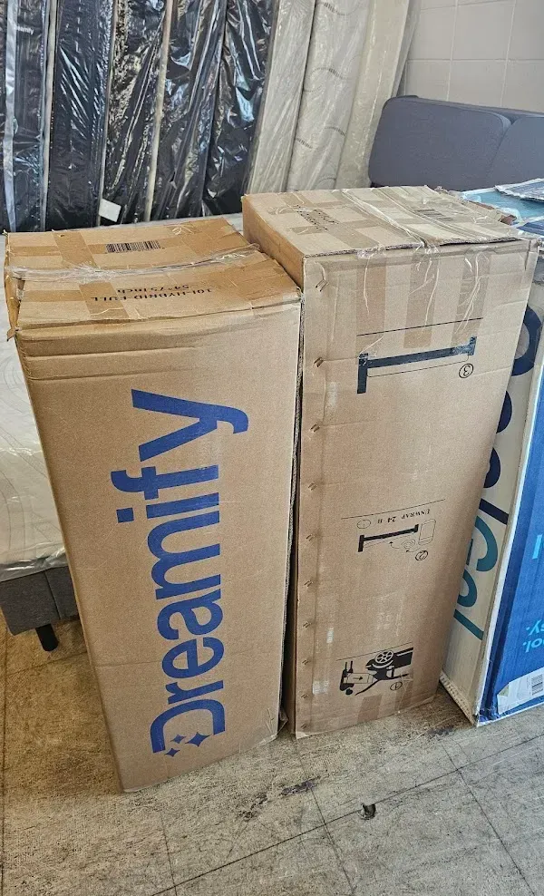 Tall cardboard shipping boxes stacked indoors, labeled “Dreamify,” with one blue bag leaning beside them.