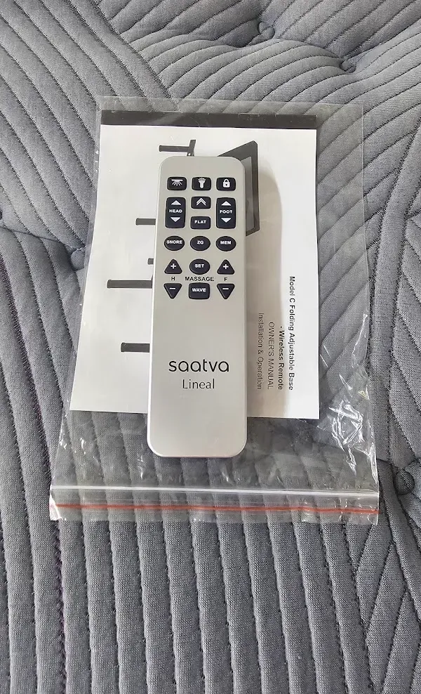 White TV remote on a gray quilted bedspread, resting on a folded paper and clear plastic bag.