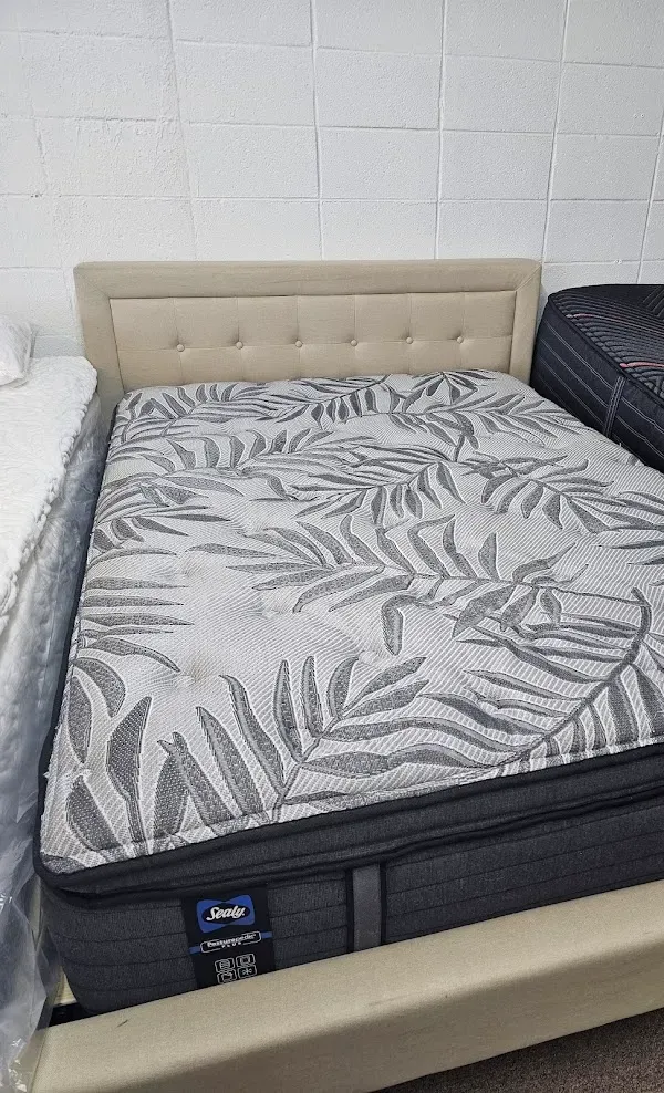 Beige upholstered bed with a gray leaf-pattern mattress cover against a white brick wall.