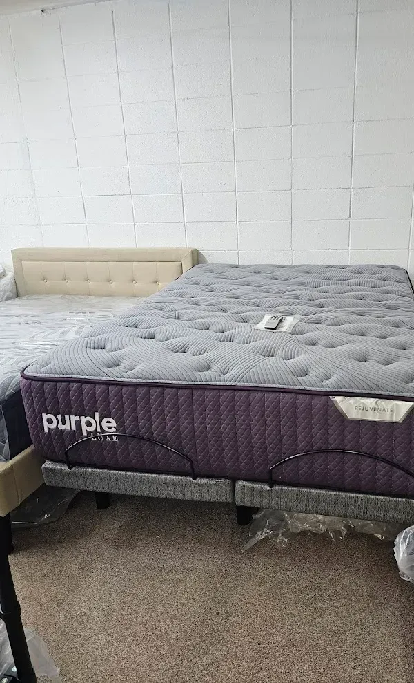 Two mattresses on bed frames against a white brick wall, one purple and one gray.