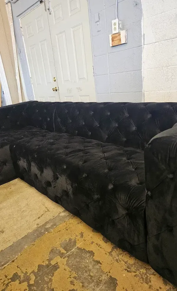 Black tufted couch against a white cinderblock wall in a worn room with a plywood floor.