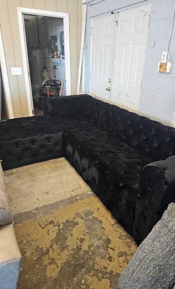 Black sectional sofa in a cluttered room with two white doors and a worn beige rug.