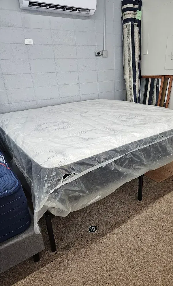 Bare mattress on a bed frame in a small room, covered with clear plastic.
