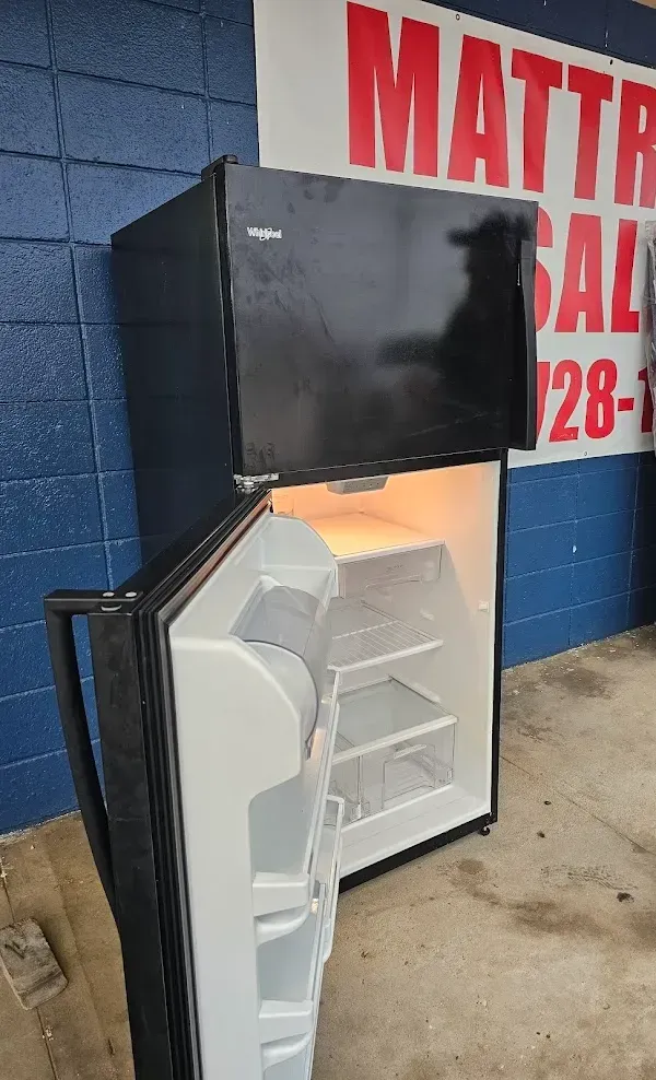 Small black mini fridge with open white door beside a mattress sale sign on a blue wall.