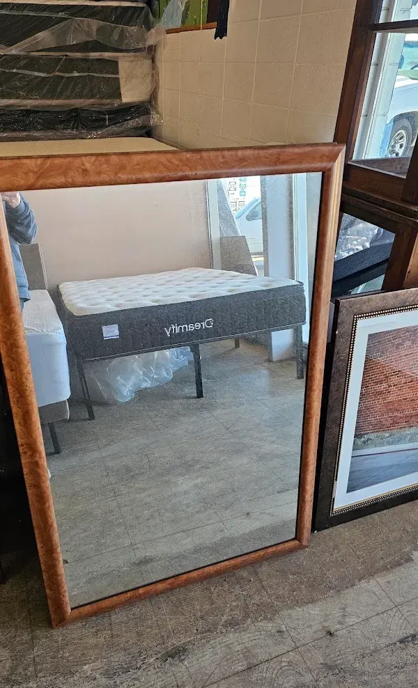 Wood-framed mirror leaning against a wall, reflecting a gray bench in a room with wood floors.