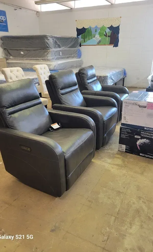 Three black recliner chairs in a store display room with stacked furniture and boxes nearby