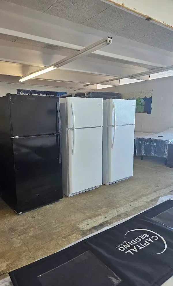 Three refrigerators in a warehouse, one black and two white, with shelving and a concrete floor.