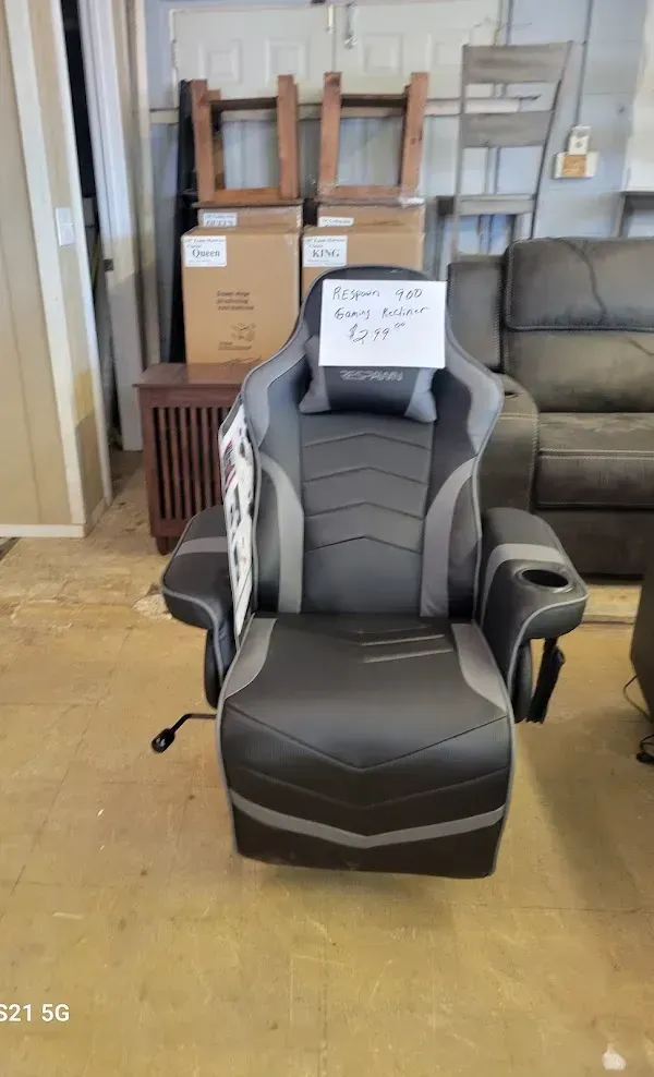 Black and gray gaming chair in a storage room with stacked boxes and a couch in the background