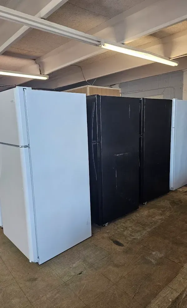 Three upright refrigerators, two black and one white, standing in a warehouse-like room