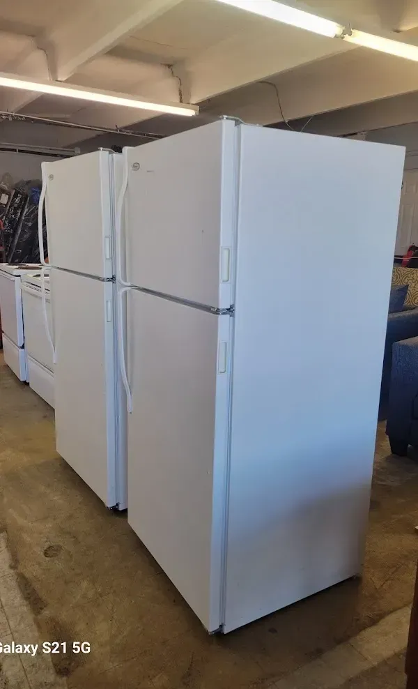 Several white refrigerators lined up in a warehouse or storage area under fluorescent lights