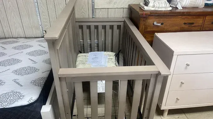 Gray crib between a bed and dresser in a nursery or bedroom, with folded bedding inside.