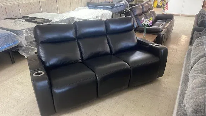 Black leather reclining sofa in a furniture showroom, with other sofas and wrapped mattresses in the background