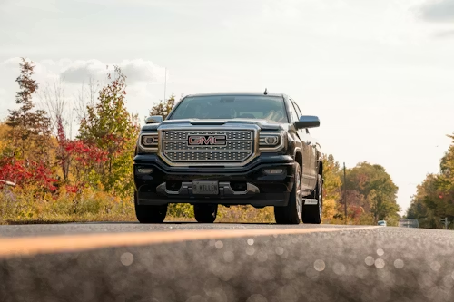 A dark-colored GMC pickup truck driving on an asphalt road surrounded by autumn trees.