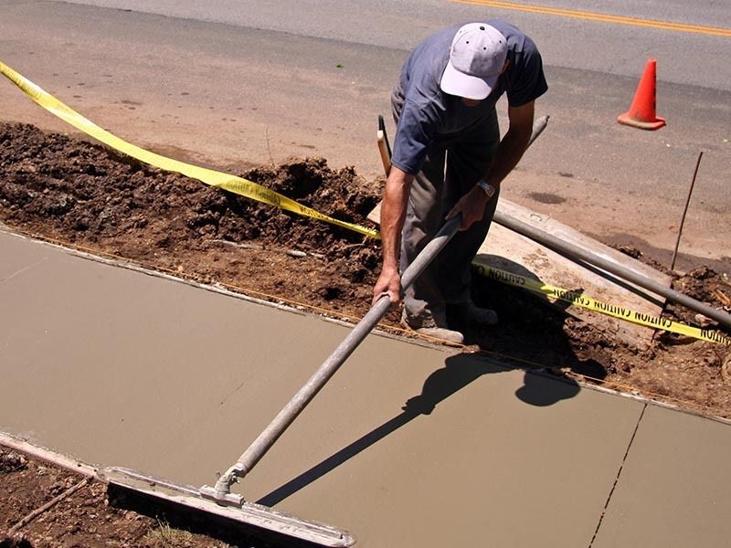 A construction worker in a white cap uses a long-handled concrete float to smooth a newly poured sidewalk.