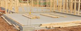 Wooden wall framing stands on a newly poured concrete foundation at a construction site.