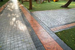 A paved walkway with light grey, dark grey, and orange stone sections, bordering a grassy area with trees.