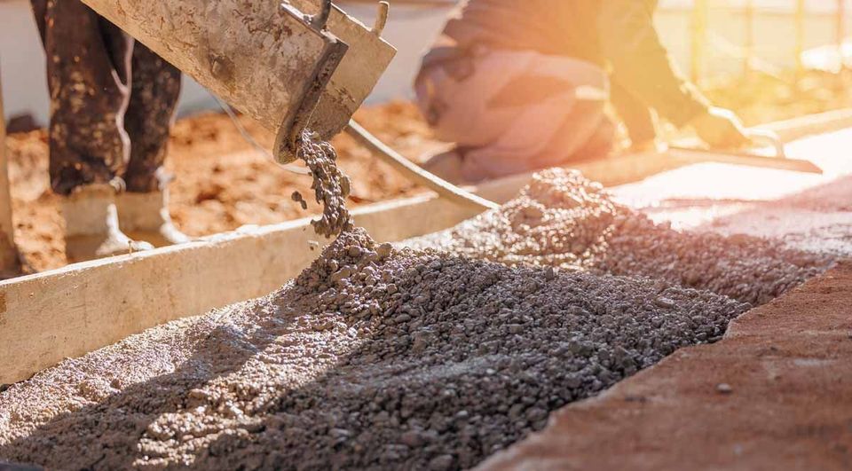 Fresh concrete pours from a chute into a wooden frame at a construction site while a worker levels the surface nearby.
