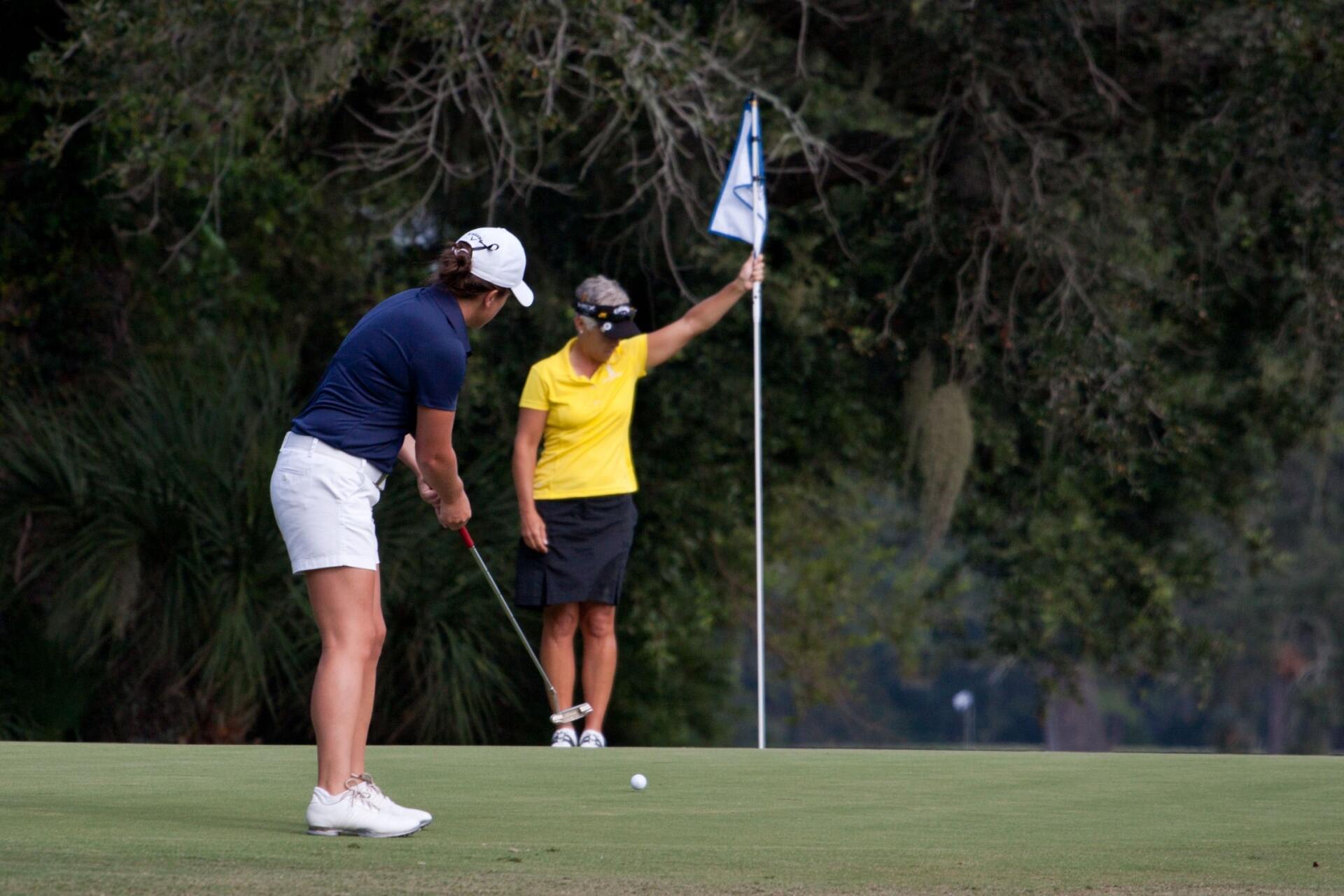 Golfer putting on green, flag tender holding flag. Course set in a wooded area.