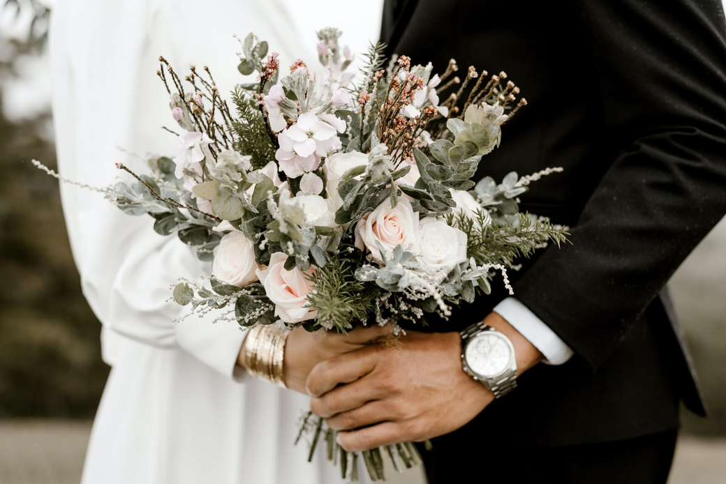 Couple holding a bouquet of light pink and green flowers, wearing wedding attire.