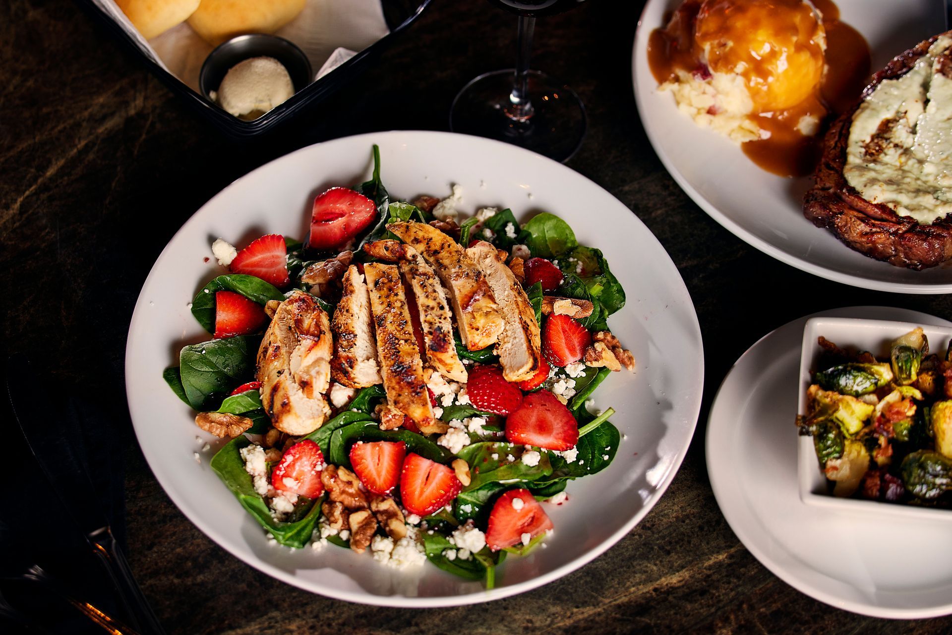 A strawberry chicken salad with feta, nuts, and greens sits next to a plate with mashed potatoes and a side of vegetables.