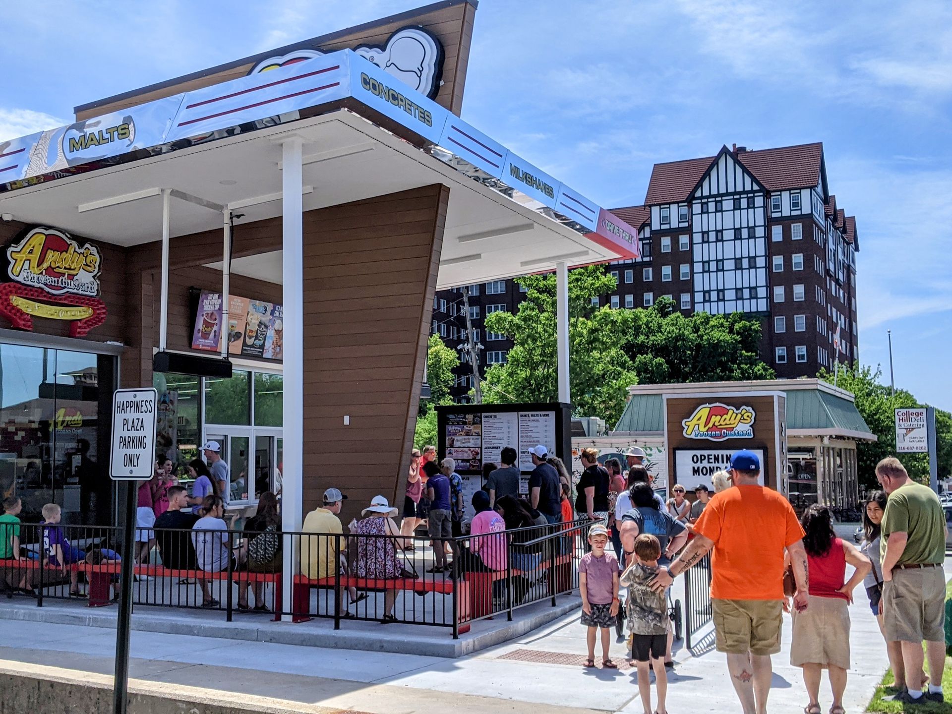 A sunny outdoor scene shows people gathered at a modern drive-in restaurant with a large, white, angled canopy roof.